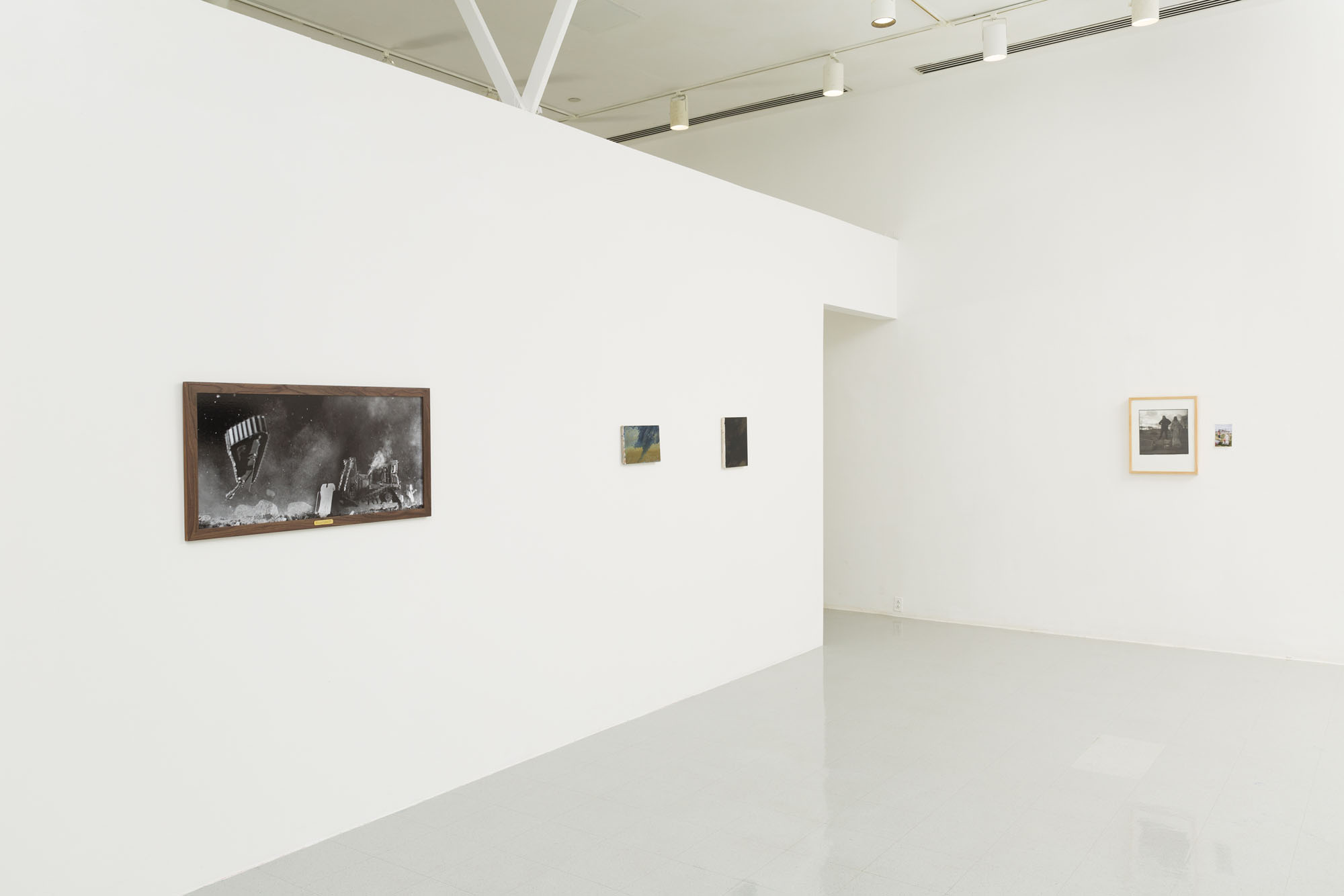 An angled view of a white wall gallery with a photograph of an exploding IDF bulldozer, two small abstract paintings in black and blues and a small framed photograph of a figure next to a pinned 4x6 photograph