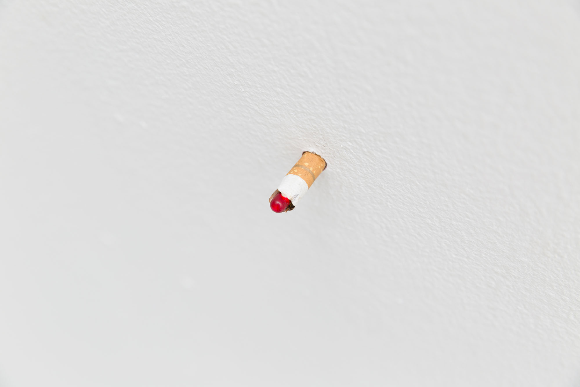 A close up view of a cigarette butt emerging from a white wall with a small red light at the end. 