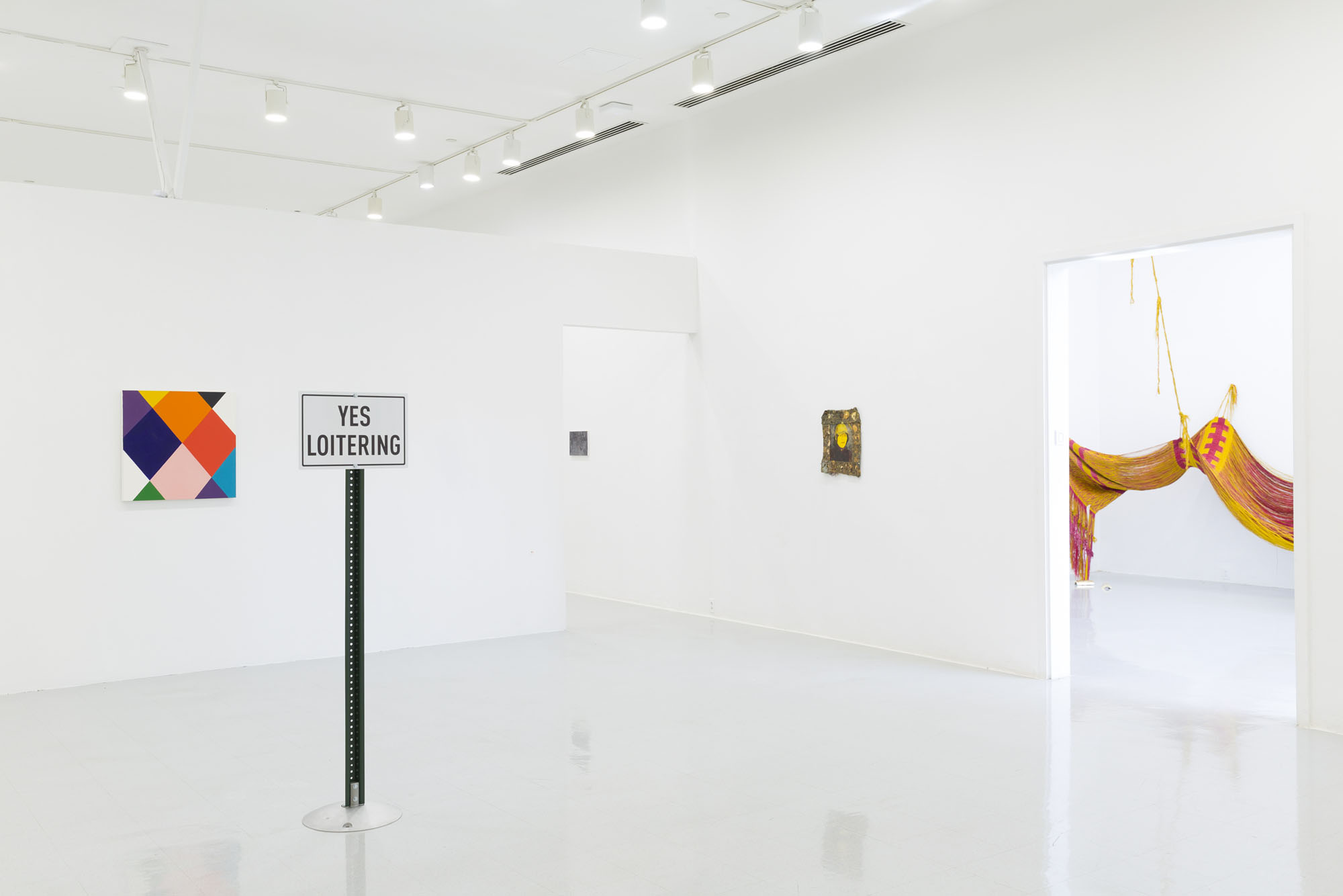 An angled view of a white wall gallery with a sign post that reads “Yes Loitering”, three paintings- a diamond plaid color field, a yellow self portrait, and through the doorway a small brown canvas. In the other doorway is the view of a large yellow and pink textile installation.