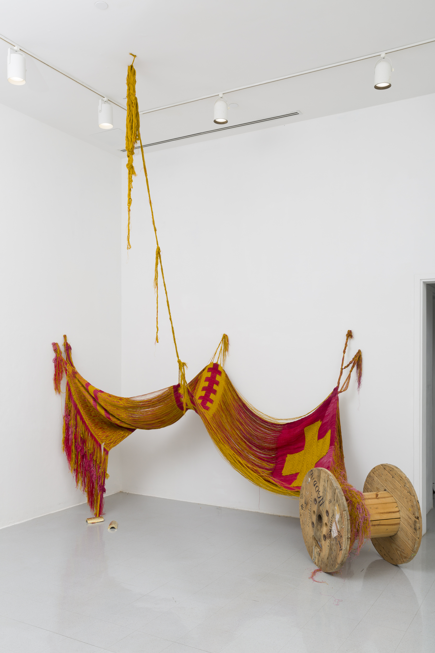 A white wall gallery space with a large scale double-woven textile installed in its corner. The textile is yellow and red-pink connected by its fringe to the walls and ceiling. On the right of the work is a symmetrical cross icon in yellow surrounded by red-pink. Its fringe cascading onto a large wooden cable reel.