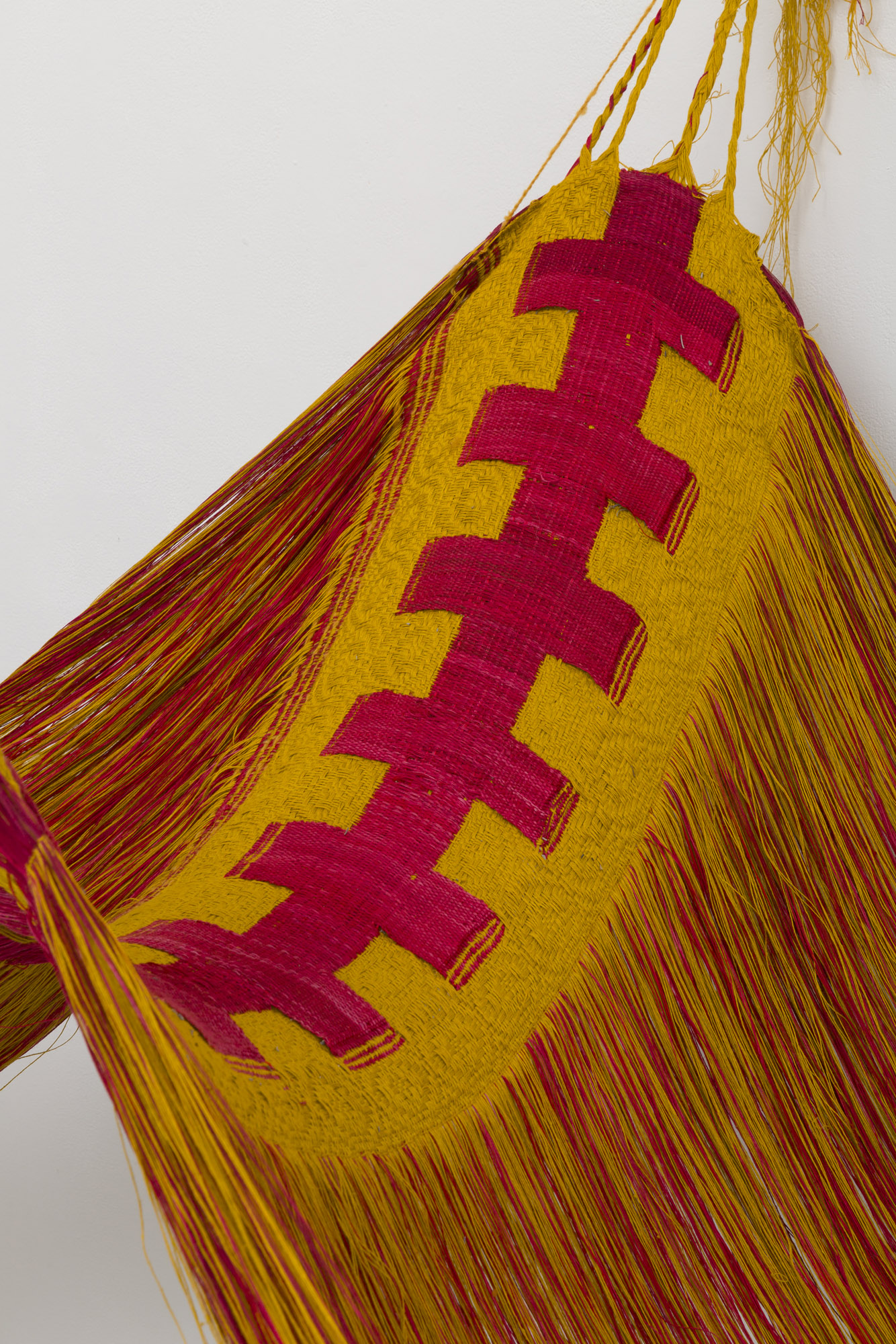 Close up of a double woven linen textile with circular patterns in golden yellow yarn surrounding a row of connected pink-red crosses which form together to look like one large surgical stitch. The textile droops in a curve and its yellow and pink fringe falls on each side