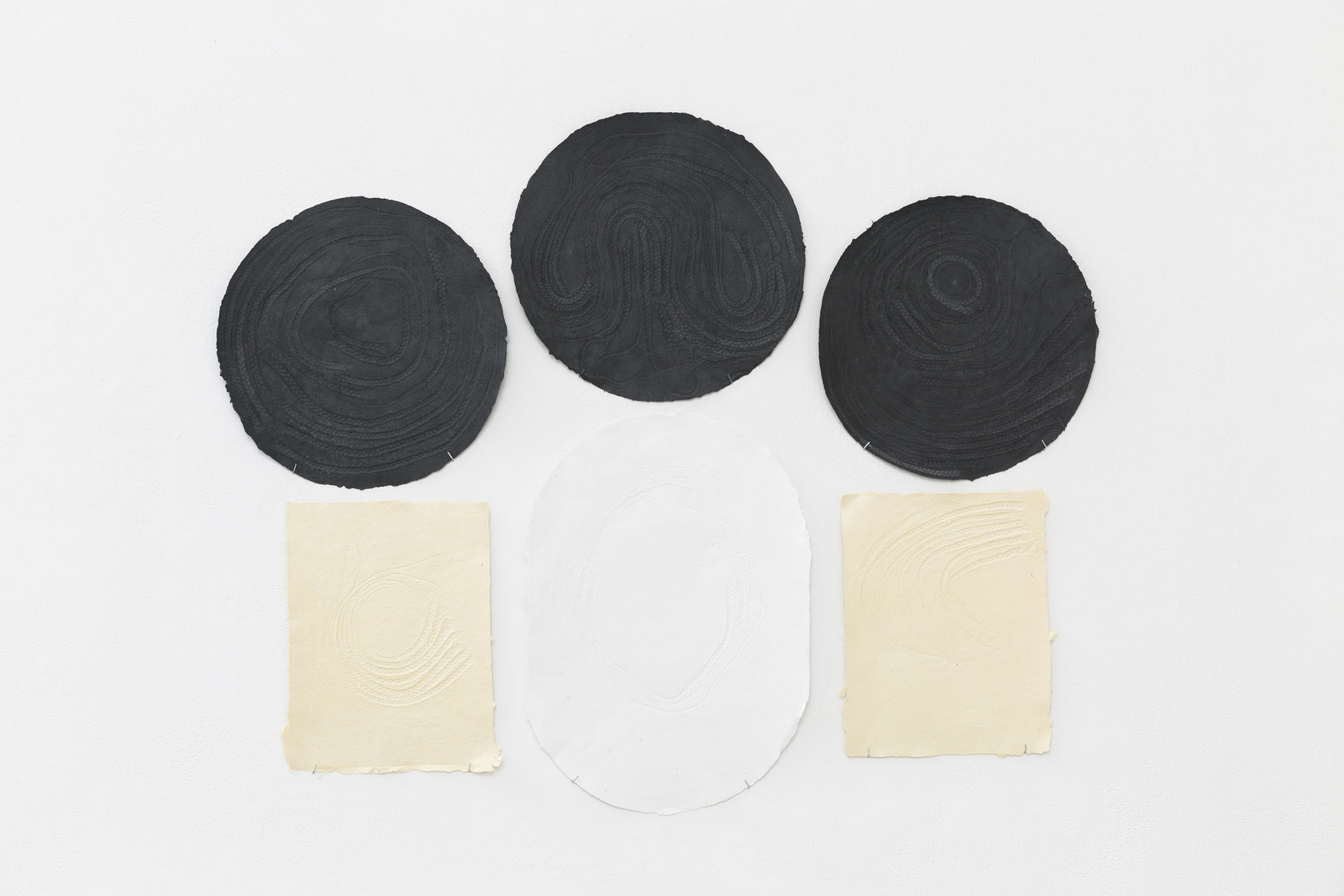 An installation of several individual handmade papers with inlaid textures made with the artist’s personal hair braids. There are three in a row: white, cream, white and above each of these is a circular black paper, each with different braid patterns.