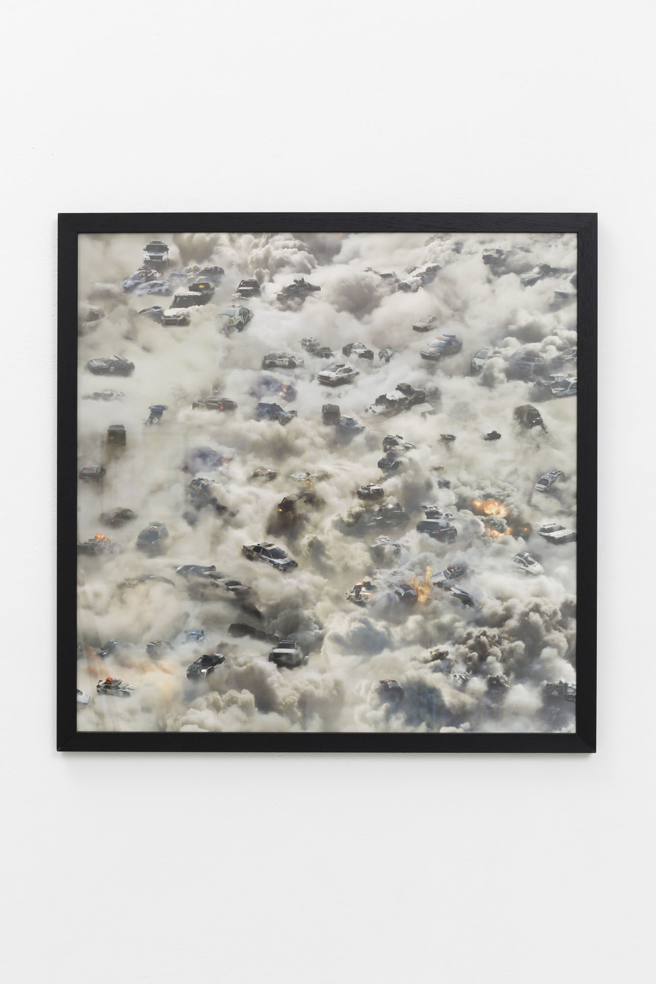 A black frame contains a print of a field of smoke with dispersed cars in various states of distress and engulfed in flames. The depth of perspective affirms clearly that this artwork was generated through Artificial Intelligence.