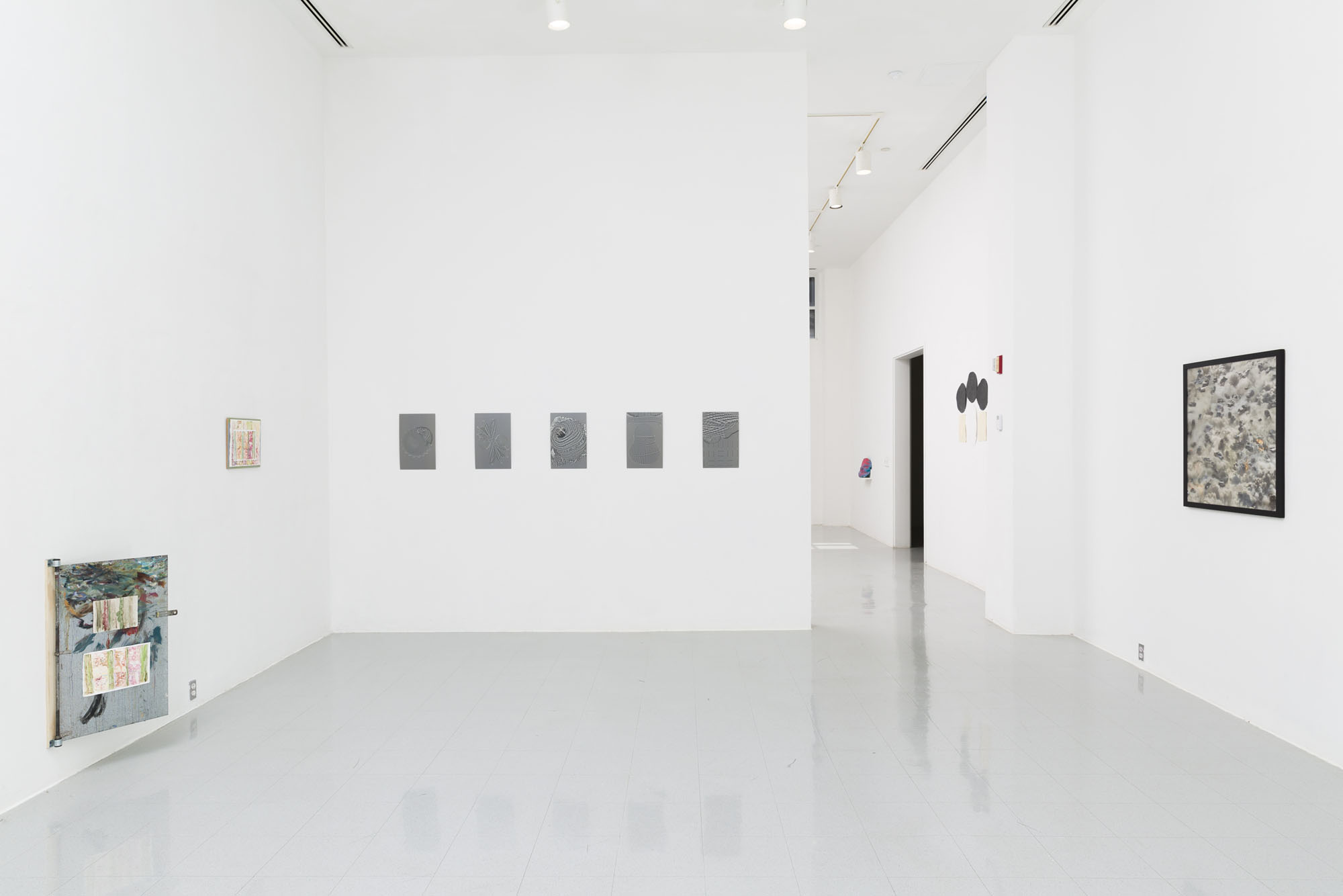 A white wall gallery with a small painted metal door the size of a painting cabinet next to another watercolor painting. On the adjacent wall are five gray 11 x 17 panels and on the final wall is a black framed print. Through the doorway two other artworks are visible.