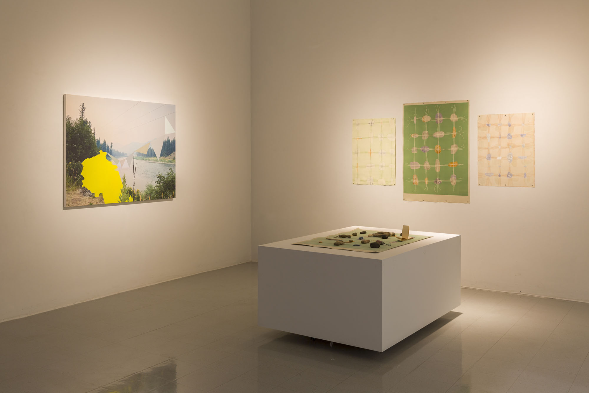 A dim-lit white wall gallery with a painting on the left of a body of water and trees with triangular flags in the foreground. On the right there is a plinth with a print on top of it and three prints on the adjacent wall with rocks strung together in grid formations