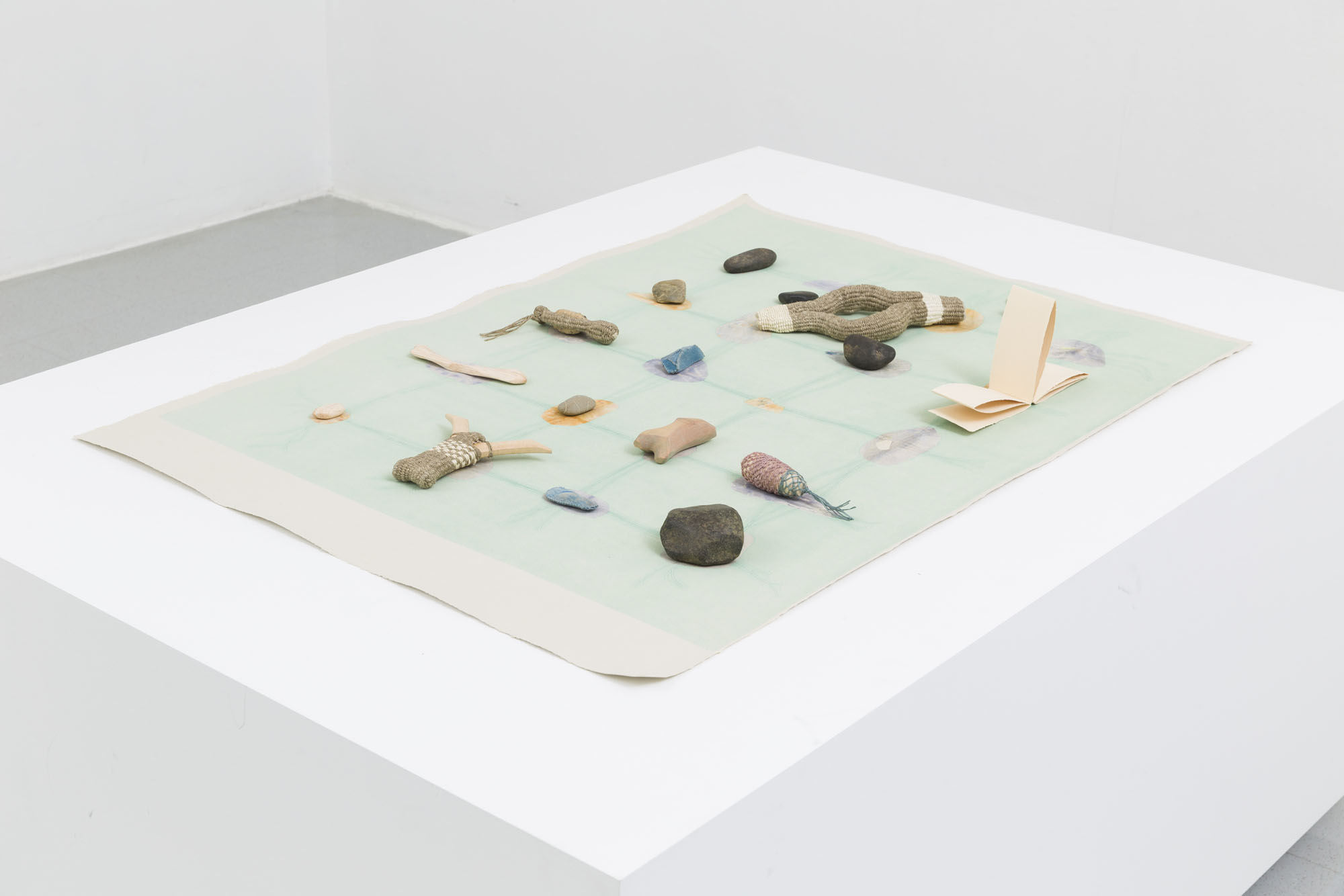 A short and wide white plinth has a print with small objects on top of it. The print has a green background with rocks strung together in a grid on its foreground. On top of this print sits several rocks, handmade baskets and a small booklet with braille embossed textures.