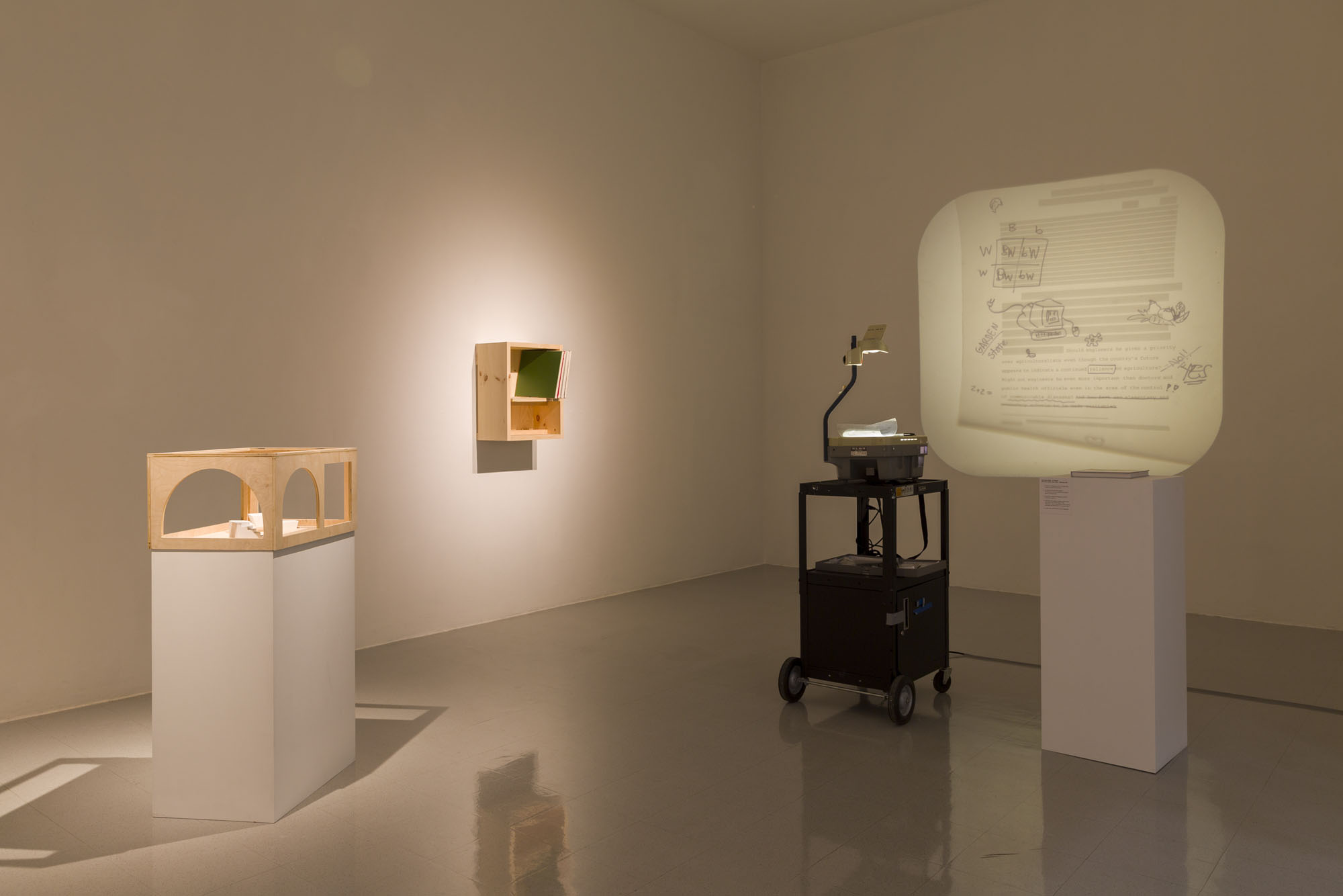 A dim lit white wall gallery. On the left is a wooden box with arch windows on top of a pedestal. There is a wood box installed on the wall with a green painting stacked inside of it. On the right is an overhead projector on a black rolling cart and a book atop another pedestal.