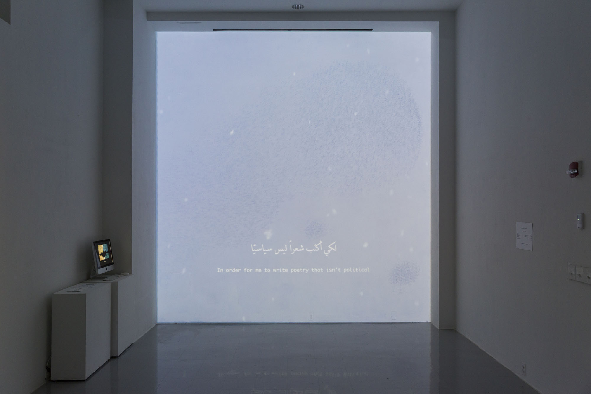 Projected video on a large wall of a starling bird pattern overlaid with white paper leaflets falling from the sky. The captions on the video read “In order for me to write poetry that isn’t political