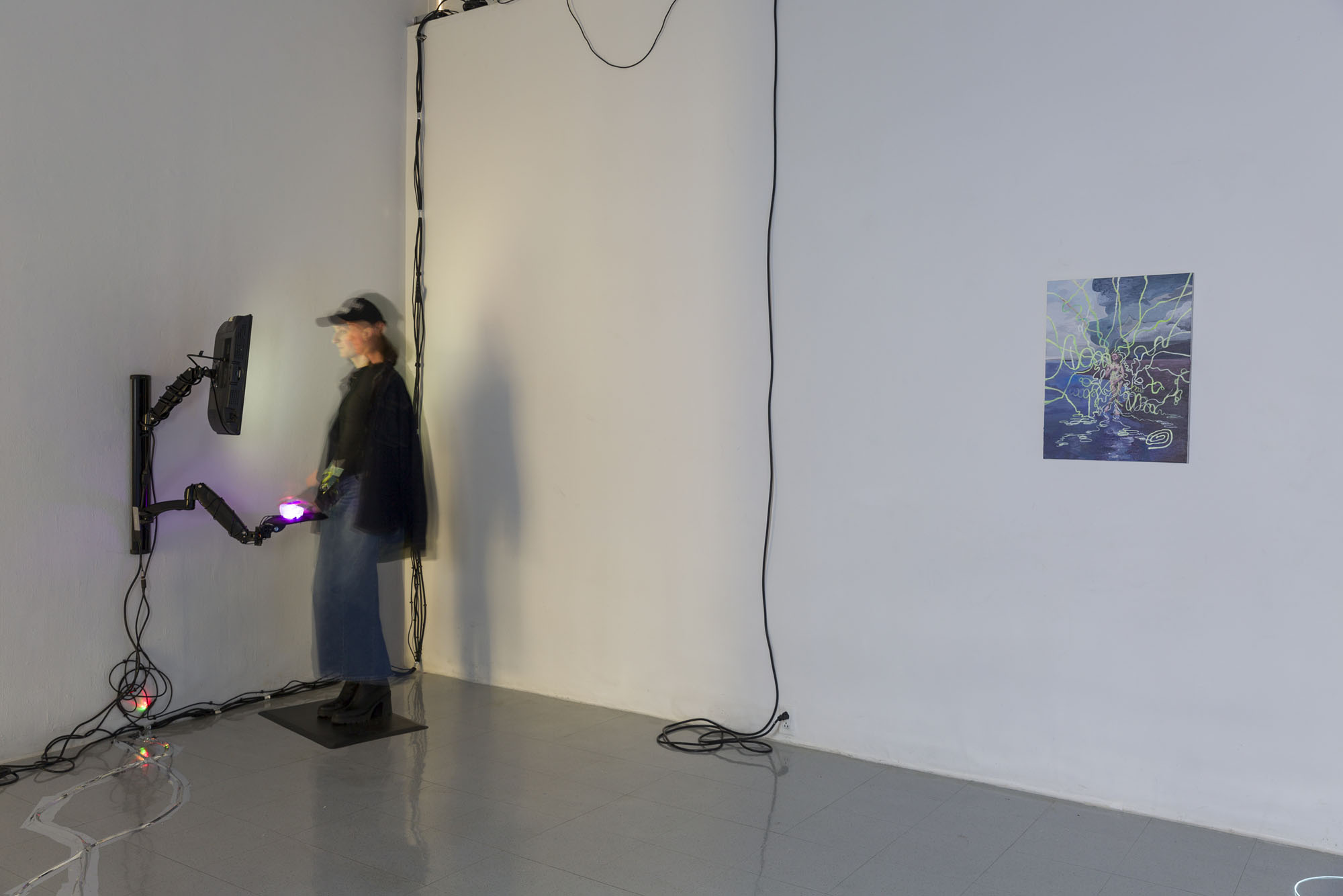 A dim lit white wall gallery. On the left Quin stands on a black anti-fatigue mat in front of a TV screen installed vertically with a stand below it with an illuminating computer mouse in pink. There are black and silver cords connecting these devices to the wall and floor. To the very right of the room is a sixteen by twenty four inch vertical painting of Quin as the Birth of Venus on a shoreline with glowy strings flowing out from her body in every direction. 