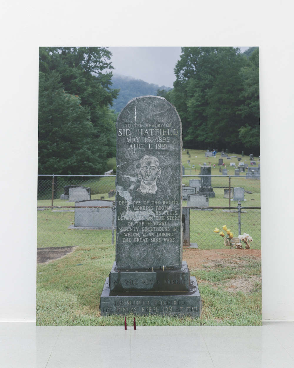 A large photograph of a tombstone in a verdant cemetery leans against a white wall. In front of the tombstone are two red training bullets standing upright.