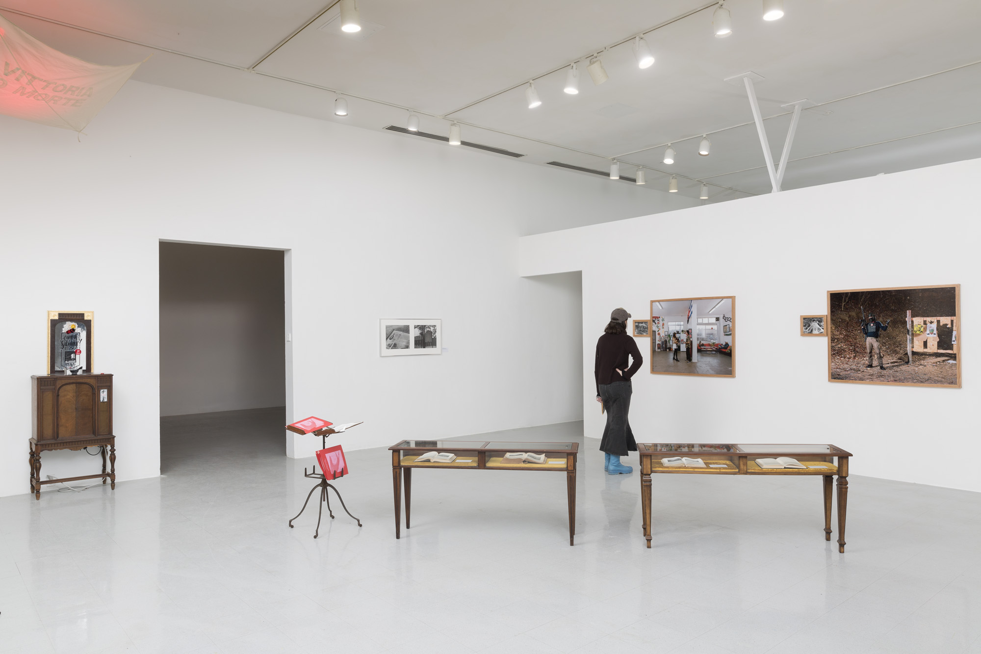 A wide view of a white wall gallery with framed photographs on the wall, antique wooden vitrines in the center of the room next to an antique book stand with a red folder on it, and a flag hanging in the top left corner above a wooden cabinet with a mirrored sign above it. A figure in all black with blue boots stands looking at one of the photographs.