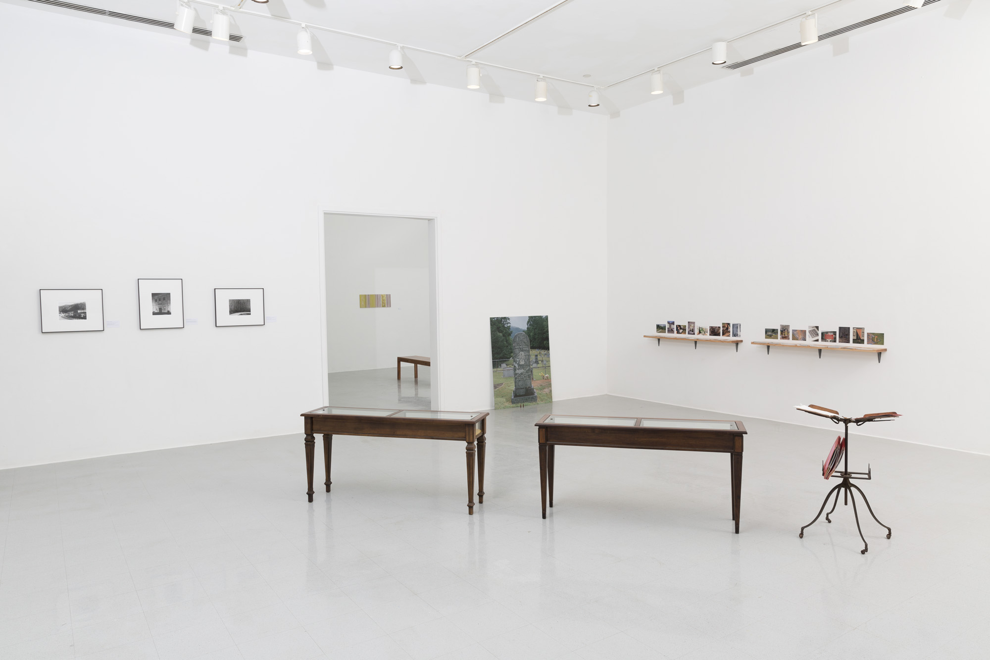 An angled view of a white wall gallery with three black and white photographs in black frames on the left wall next to a doorway. Two antique wooden vitrines and an antique book stand are seen from behind in the middle of the room. A photograph of a tombstone leans against the wall on the other side of the doorway, around the corner from two long wooden shelves with small vinyl images on the wall above the shelves.