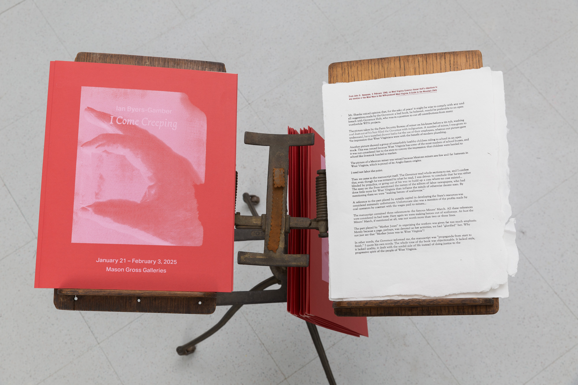 A downward looking view of an antique book stand. On the left is a red folder with a screenprinted image of a hand in white ink with the text Ian Byers-Gamber, I Come Creeping, January 21 - February 6, 2025, Mason Gross Galleries. On the right is a memo letterpressed on handmade paper.