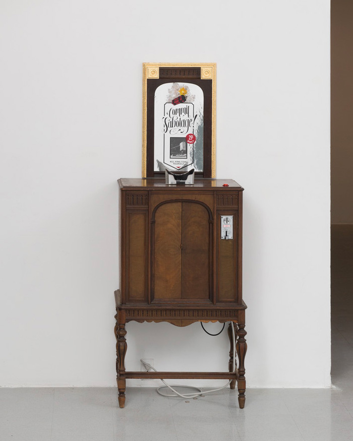 An antique wooden cabinet with an arcade machine style quarter acceptor sits against a white wall. There are power cords running down below the cabinet. On the top surface of the cabinet is an eyepiece from an old stereogram viewer. Above the cabinet is a mirrored gilded frame, on the surface of the mirror is a painted image of a bundle of dynamite with the words 