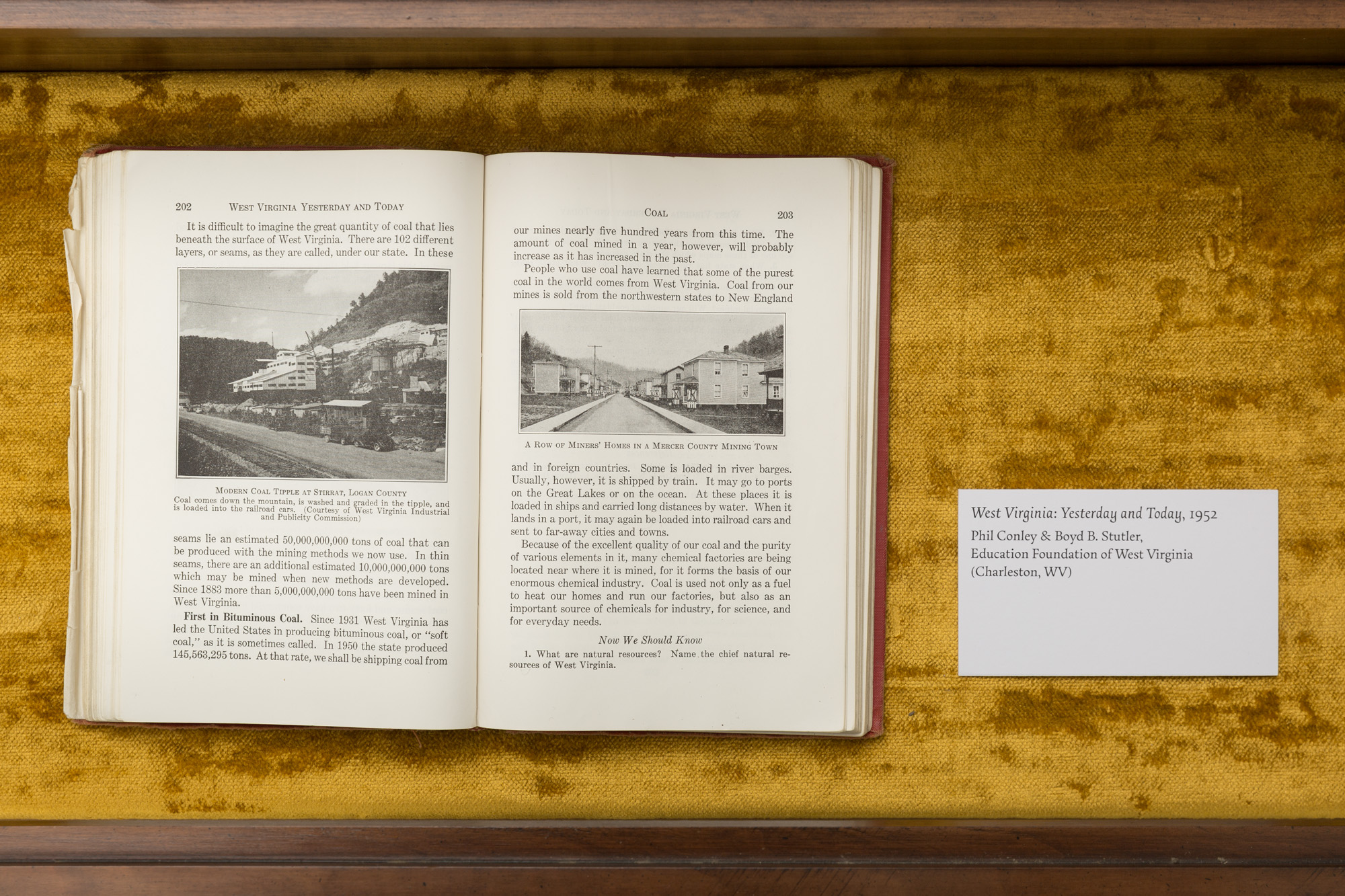 A history textbook open to a page about coal mining in West Virginia rests on a gold fabric pad inside of an antique vitrine. A label next to the book reads 