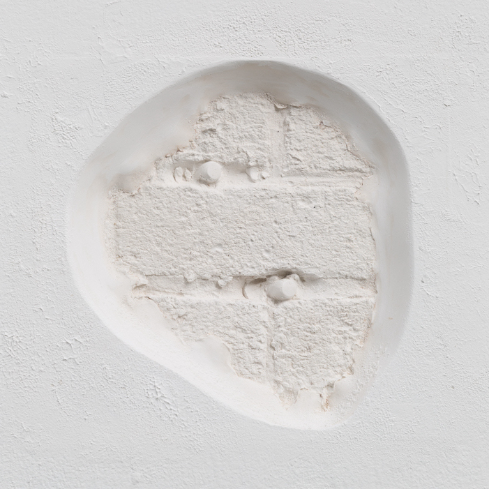 A plaster cast of a brick wall with fake bullets set into bullet holes is inset into the white wall of the gallery.