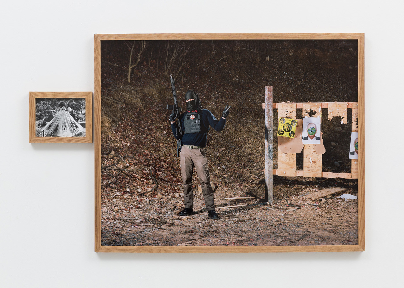 A small wooden frame containing a black and white archival image of a tent camp of striking coal miners next to a large wooden frame containing a full color image of a figure wearing a kuffiyeh and red bandana posing with a gun at a wooded shooting range. Beside the figure is a target stand full of bullet holes, two of the targets are of Richard Sackler's face. The colors of the center of his face are inverted in a circle.