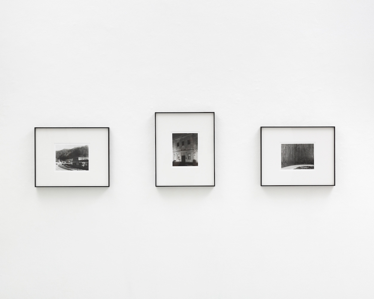 Three black and white photographs in black frames with white mats on a wall. The left photograph is a landscape image of a small town with a train running across the frame. The center image is of a stone bank building facade at night. The right image is of a dense forest with a curved road cutting across the foreground.