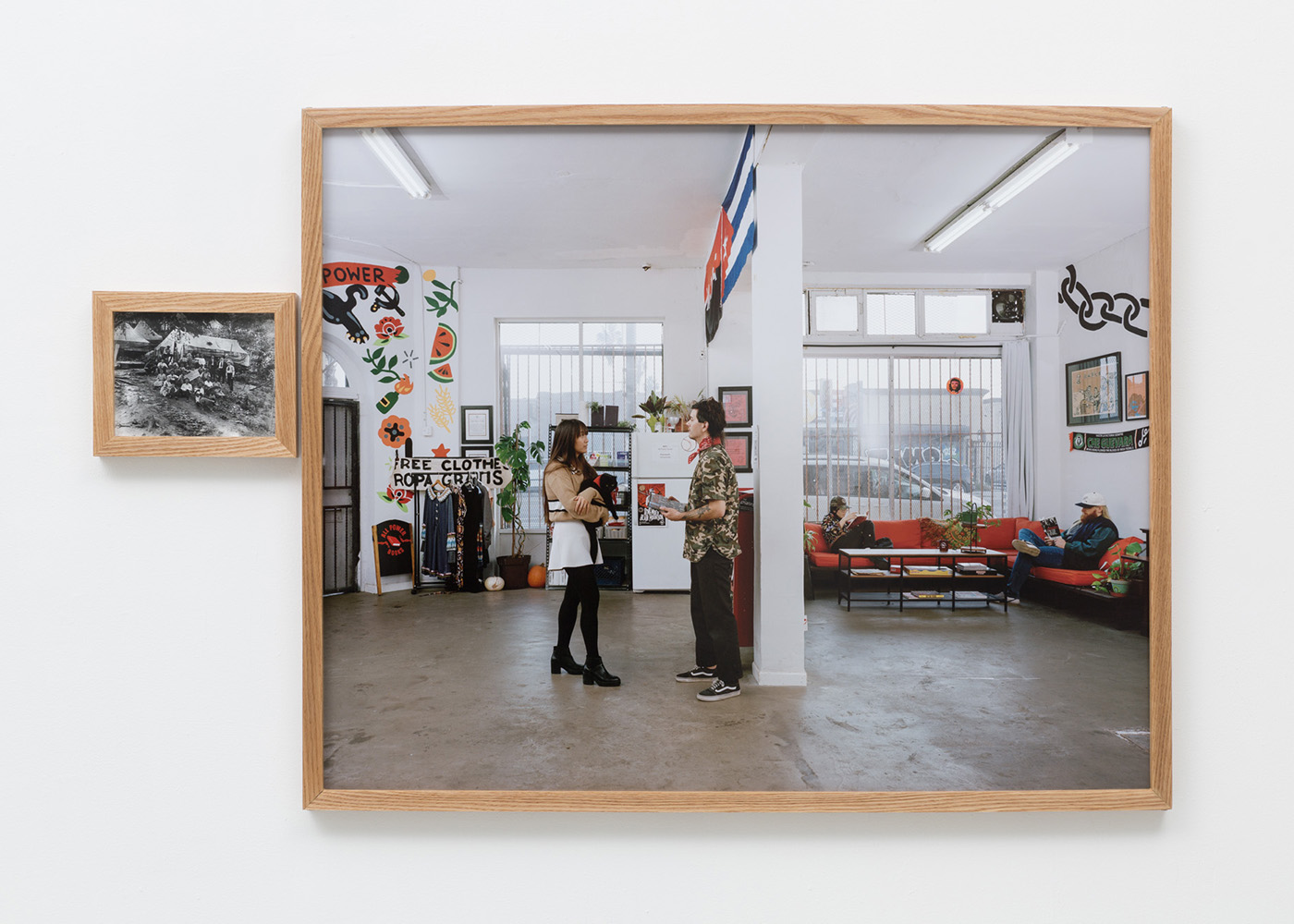 A small wooden frame containing a black and white archival image of a tent camp of striking coal miners next to a large wooden frame containing a full color image of the interior of a contemporary leftist organizing space, with people holding books and items related to the Battle of Blair Mountain.